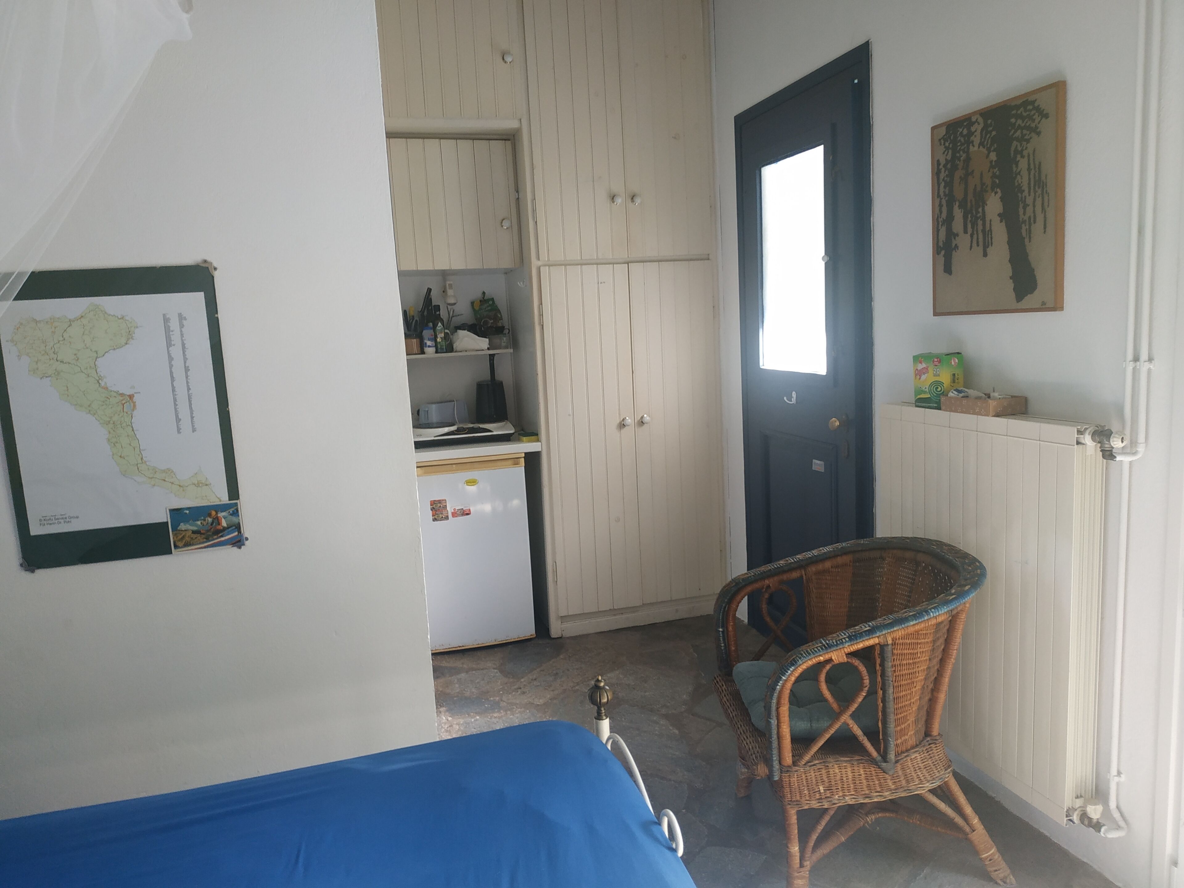 A room with a bed, a wicker chair, a map on the wall, a door, and a kitchen area with cabinets and a refrigerator.