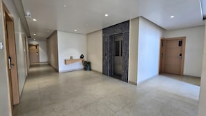 Un pasillo de hotel con piso de mármol, un estante de madera con planta en maceta, un ascensor y una puerta con placa de número.