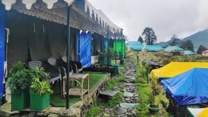 A row of tents with outdoor seating and potted plants, set in a natural landscape with mountains in the background. - RIVER SIDE CAMP CHOPTA  (Pokhri)