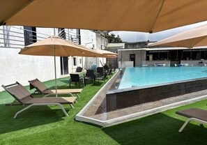 Una piscina con lettini e ombrelloni, circondata da un edificio con rivestimento in cemento.