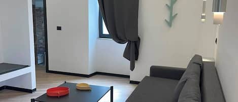 A modern living room with a sofa, a coffee table, and a TV. There is a window with curtains, a wall-mounted light, and a small table with a remote control.