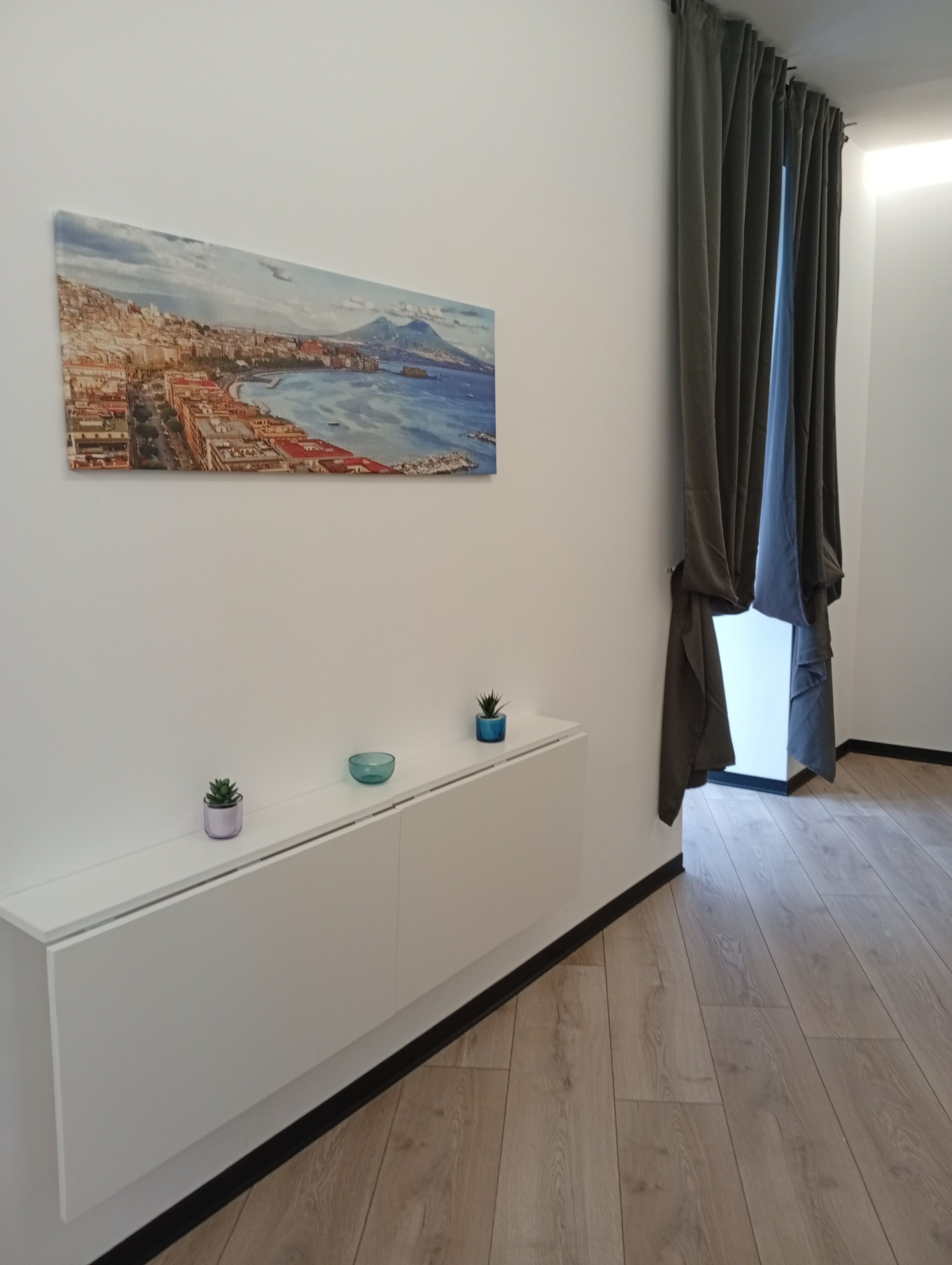 A room with a white console table, a painting of a coastal cityscape, a potted plant, and a view through a window with curtains.