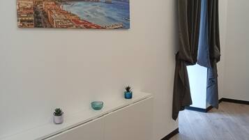 A room with a white console table, a painting of a coastal cityscape, a potted plant, and a view through a window with curtains.