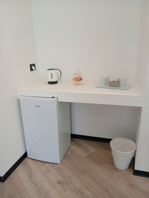 A compact kitchenette with a white refrigerator, a kettle, a small fruit jar, and a tray with cups and a basket.