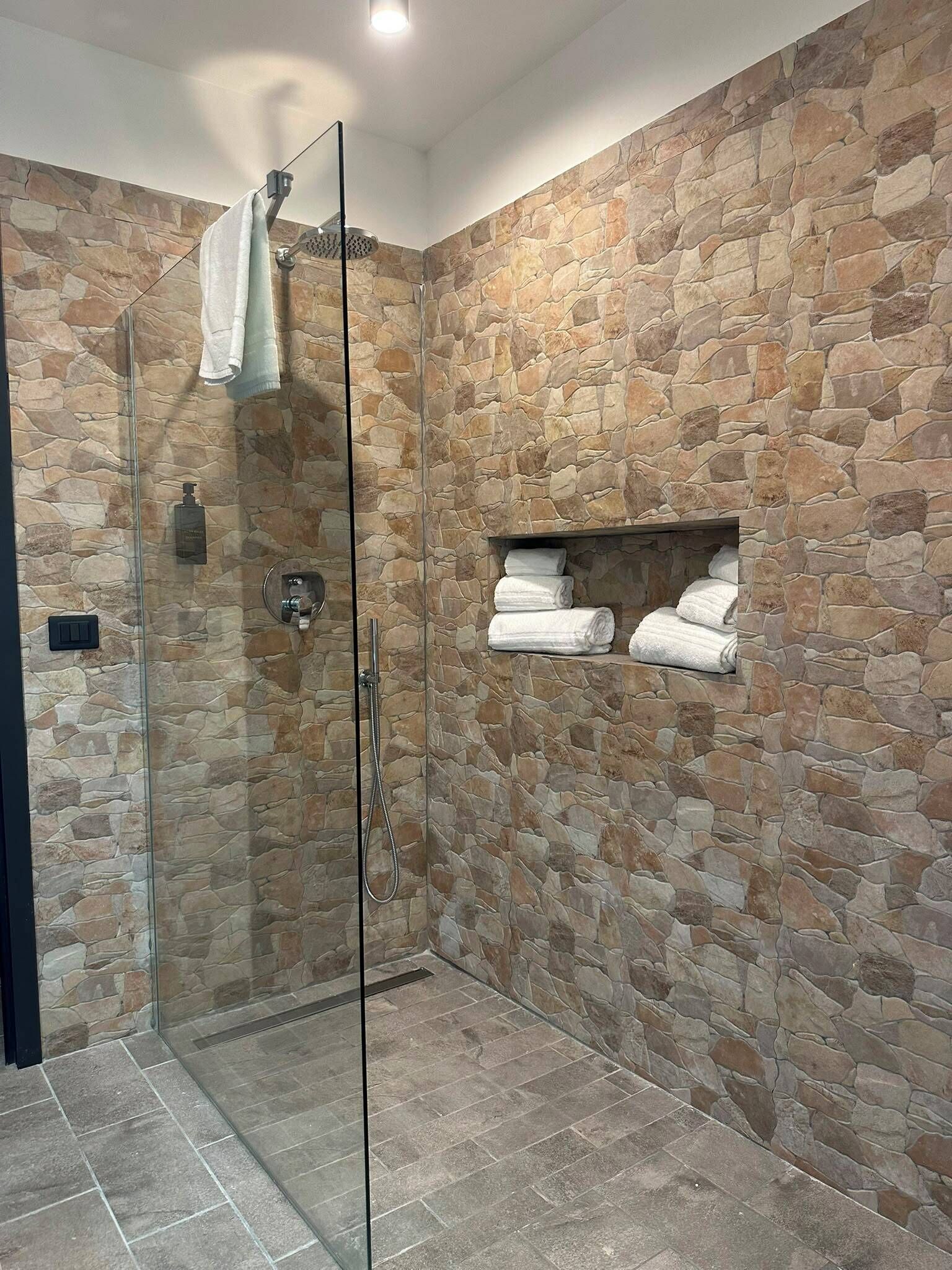 Una ducha con cerramiento de vidrio, paredes de piedra y un estante con toallas.