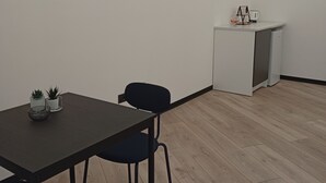 Quarto com televisão de ecrã plano na parede, kitchenette pequena com frigorífico, mesa com cadeiras e plantas em vasos.
