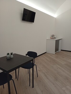 A room with a flat-screen TV mounted on the wall, a small kitchenette with a fridge, a table with chairs, and potted plants. - Borgo Double V (Napoli)
