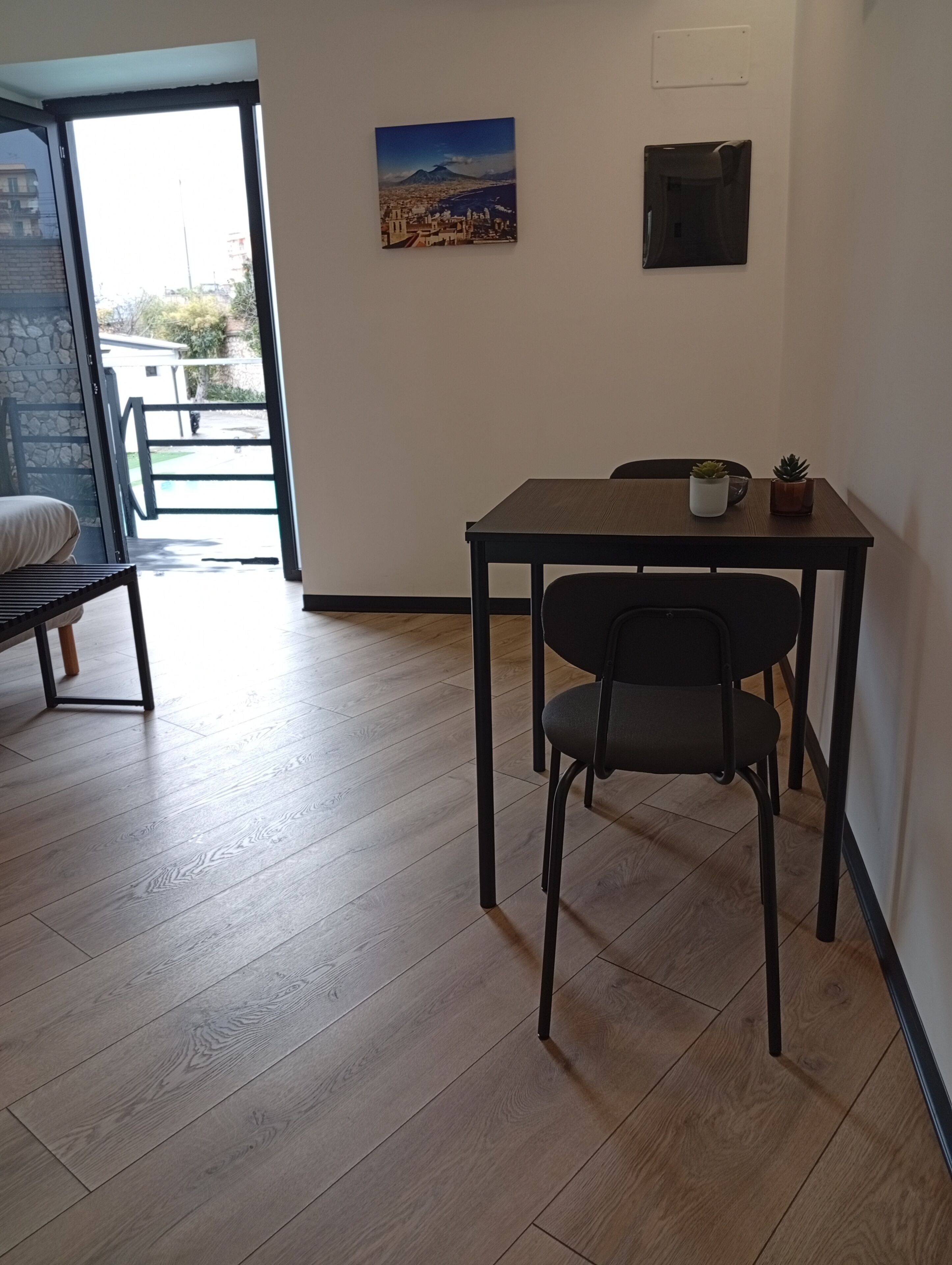 Een kamer met een houten vloer, een eettafel met twee stoelen, een kleine potplant en een aan de muur hangend schilderij.