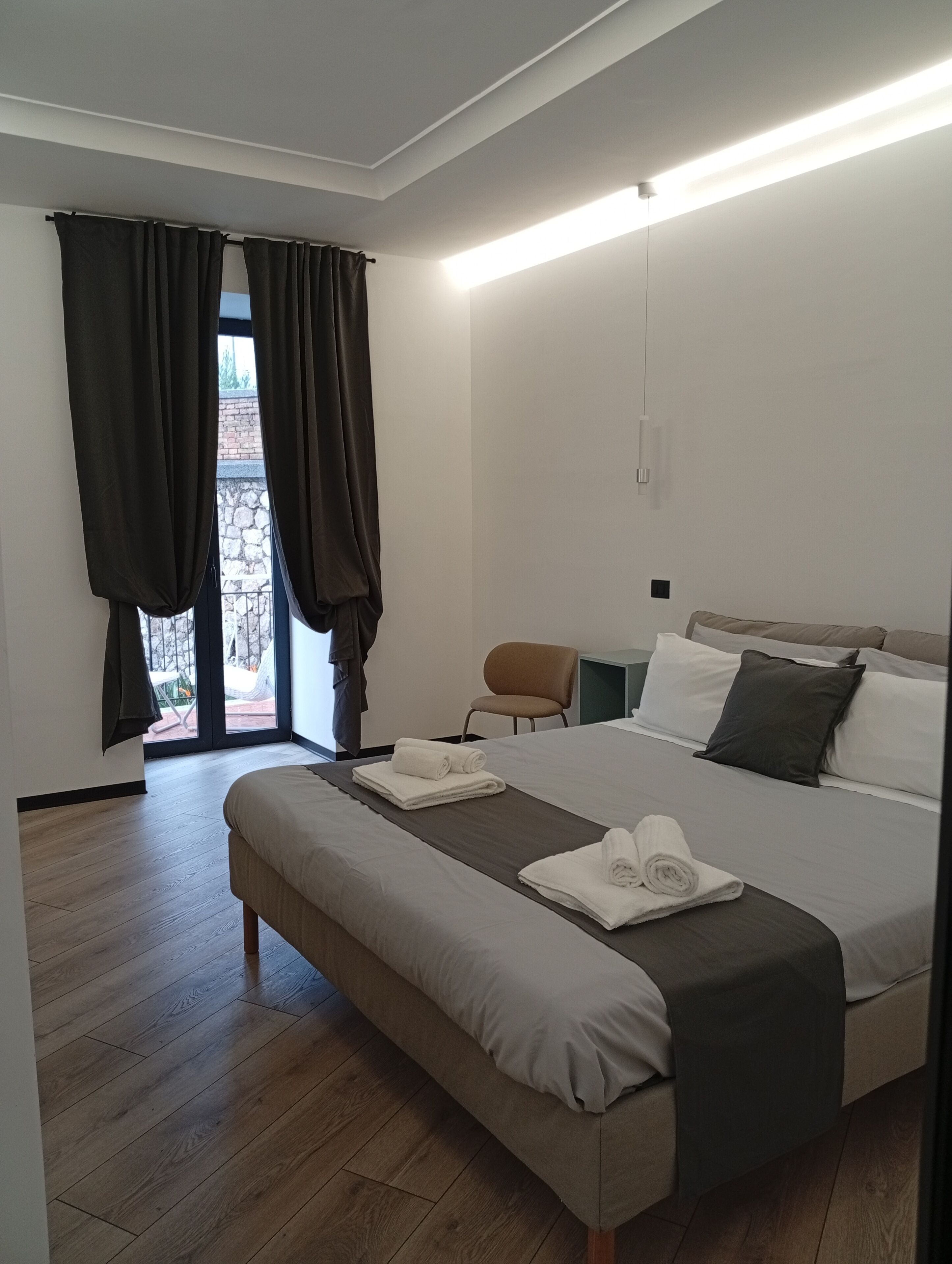 Una habitación de hotel moderna con una cama grande, un banco, una silla y vistas a través de una ventana con cortinas.