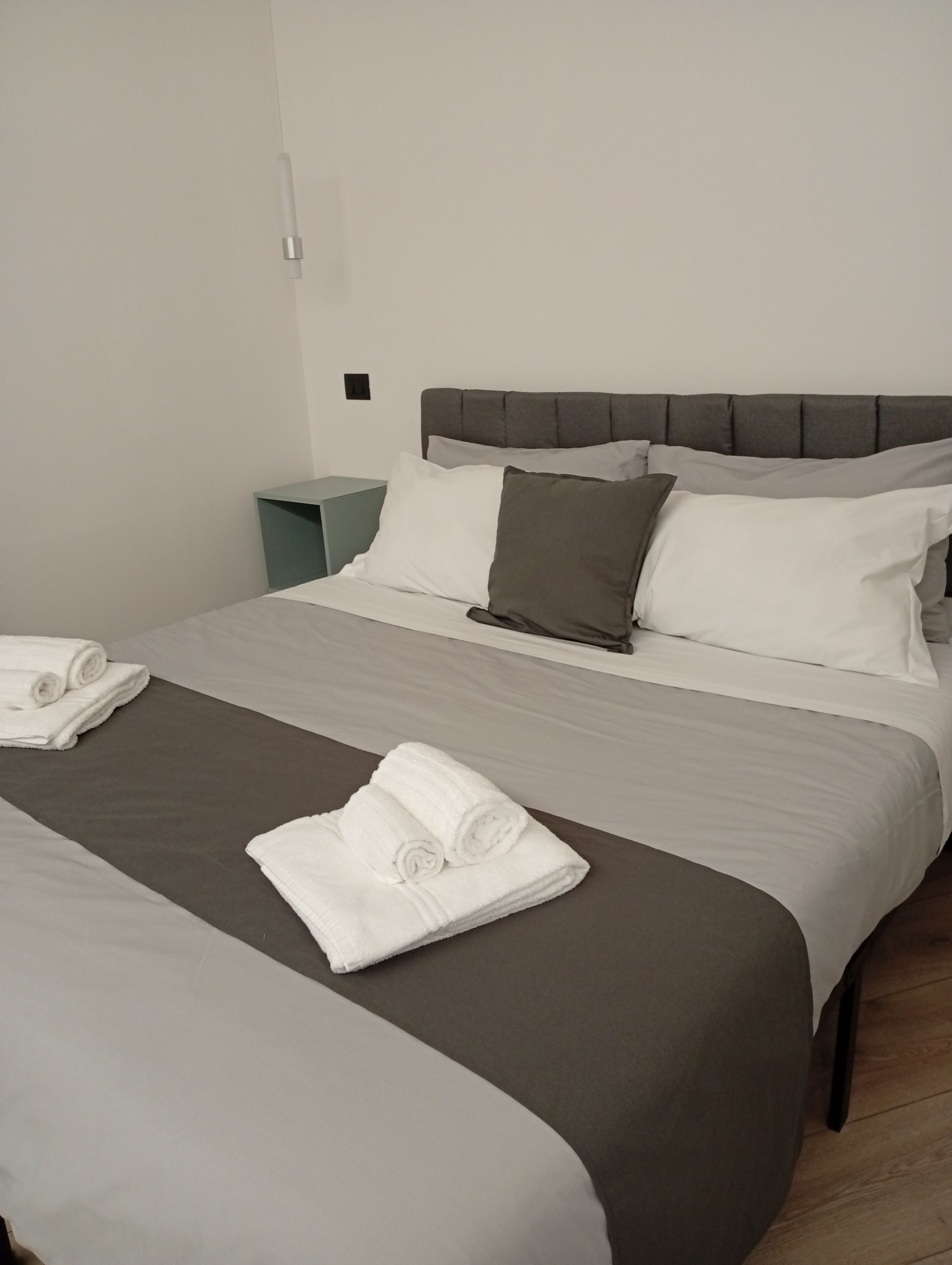 Una cama doble con cabecera gris, ropa de cama blanca y gris, y dos toallas enrolladas sobre la cama.