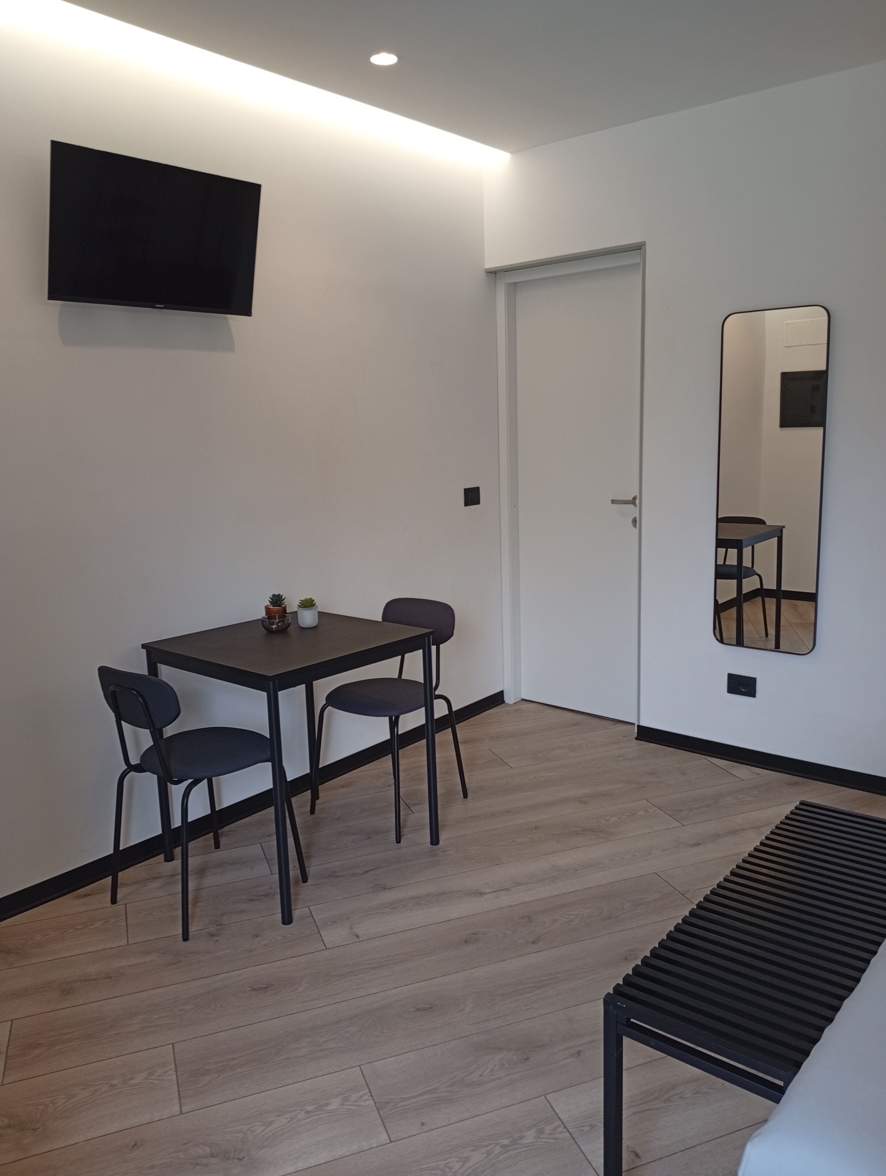 A modern room with a flat-screen TV mounted on the wall, a dining table with two chairs, a bench, and a large mirror reflecting the room.