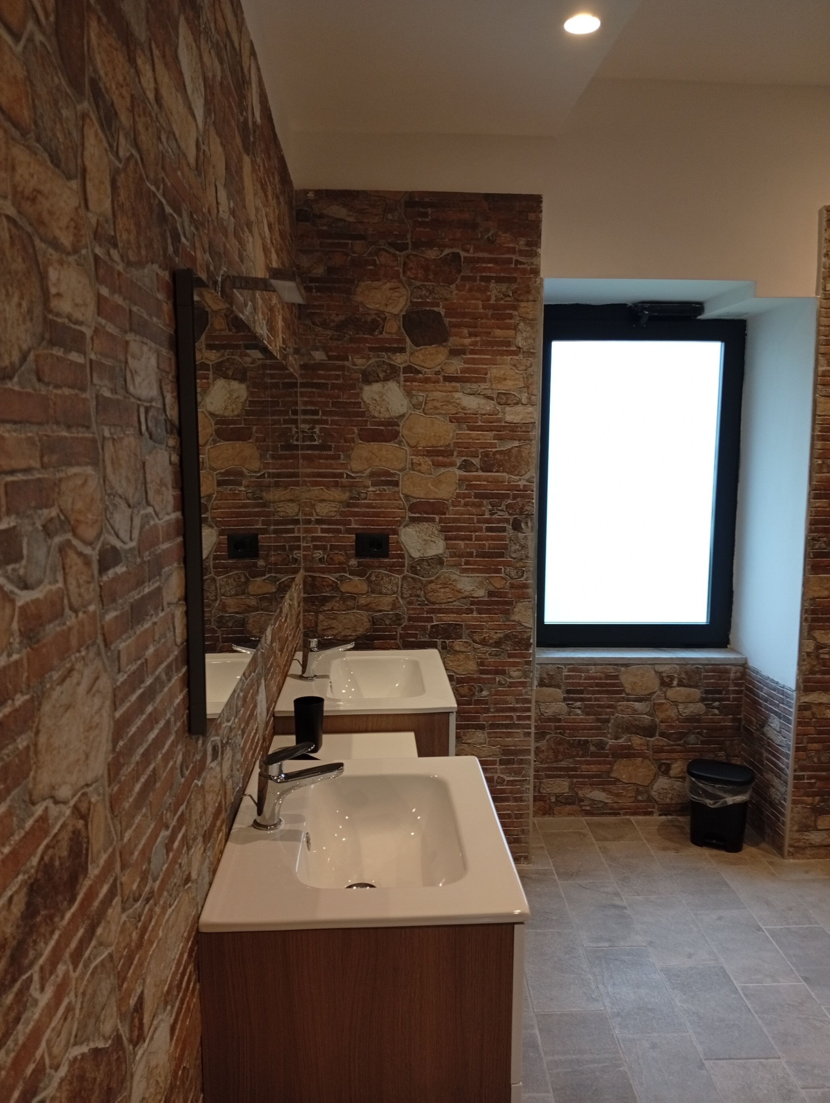 A bathroom with a stone wall, two sinks, a mirror, and a window.
