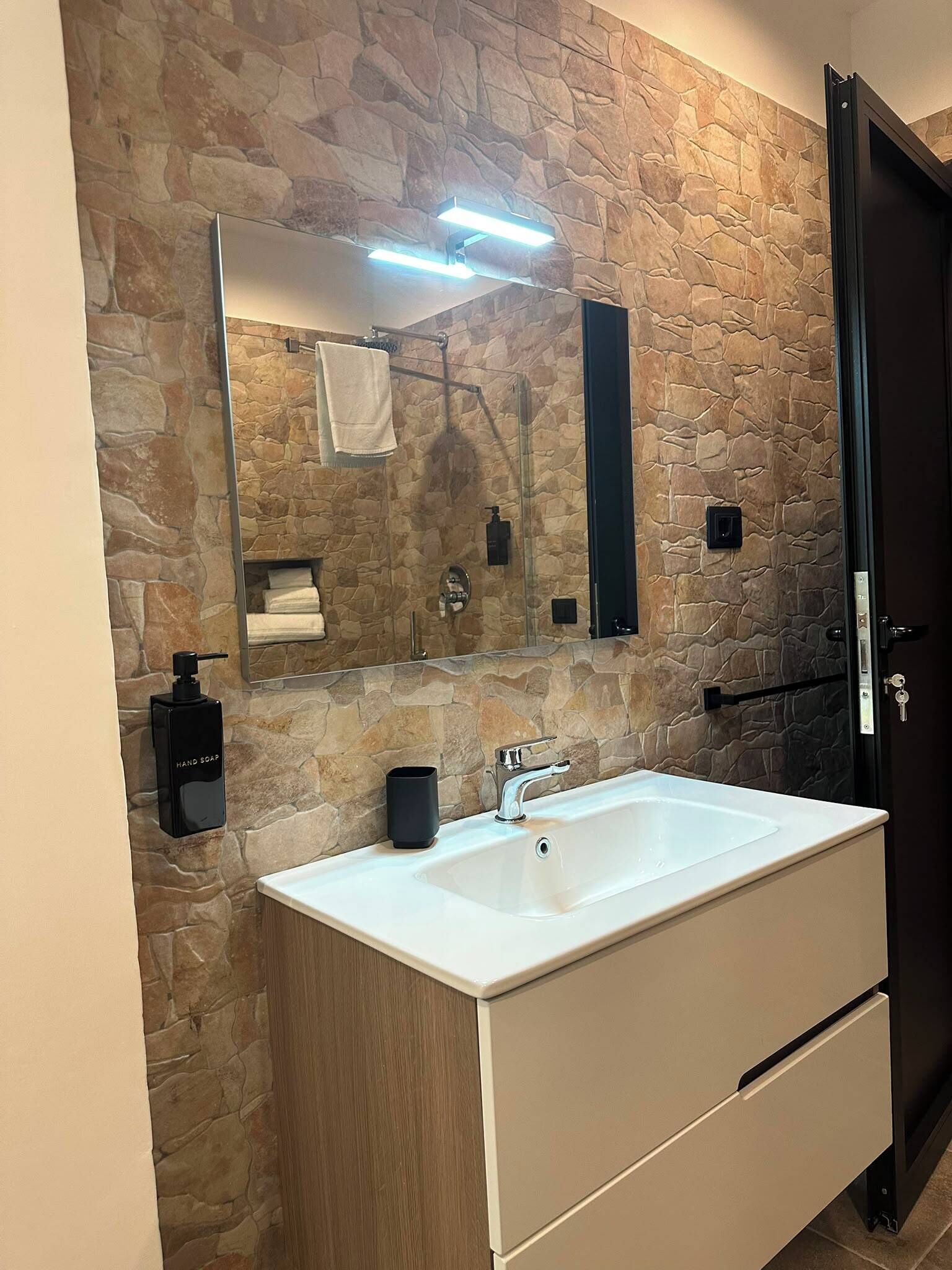 Un baño con pared de piedra, un espejo grande, un lavabo blanco, un dispensador de jabón negro y un perchero para toallas.