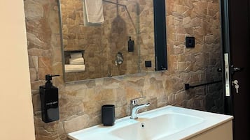 Un baño con pared de piedra, un espejo grande, un lavabo blanco, un dispensador de jabón negro y un perchero para toallas.