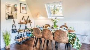 A dining area with a table set for four, surrounded by chairs, a window with a view of trees, and a shelf with various items. - Pine Needle Lodge by Interhome (Invergarry)