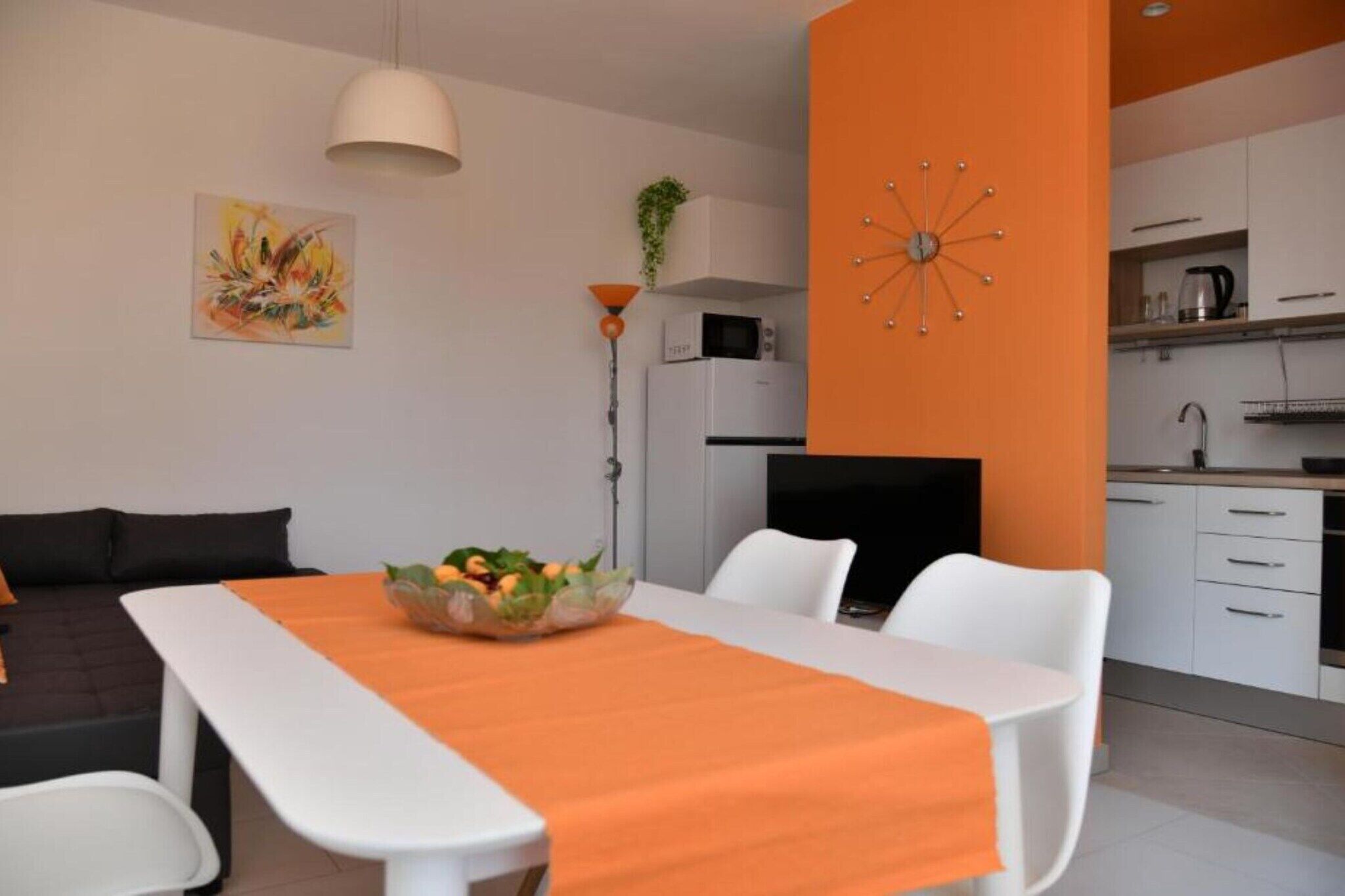 Ein modernes Wohnzimmer mit Esstisch, weißen Stühlen, einem Fernseher und einer Küchenzeile mit Schränken und Geräten.
