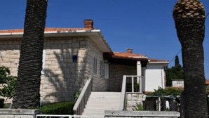A stone building with a tiled roof, a staircase leading to the entrance, and palm trees in the foreground. - One Bedroom Apartment with Terrace & Garden View (Supetar)