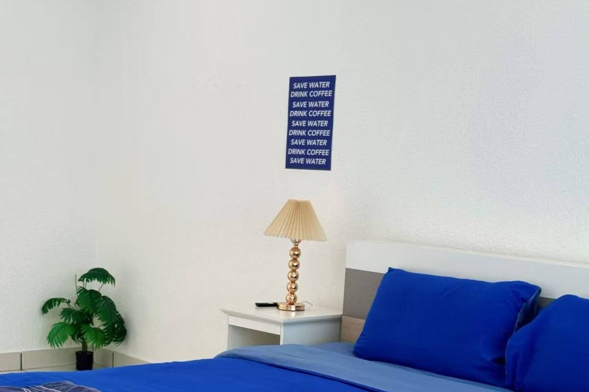 A neatly made bed with blue bedding, a bedside lamp, a potted plant, and a sign on the wall.