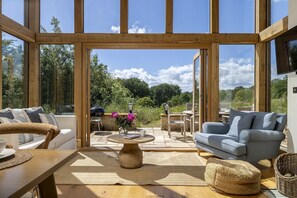A spacious room with a large window, wooden beams, a sofa, a coffee table, and a wicker ottoman. - Beautifully presented holiday home in the Cotswolds with amazing views - The Tractor Shed (Combrook)