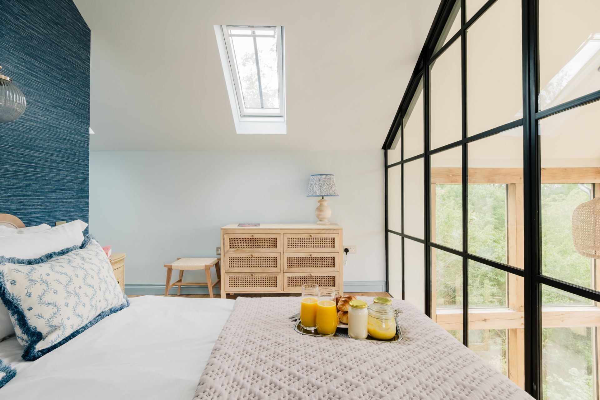 Une chambre à coucher avec un lit, une table de chevet avec une lampe, une commode et un plateau de petit-déjeuner avec du jus d’orange et des viennoiseries.