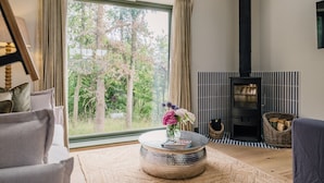 Un salon chaleureux avec une grande fenêtre, une table basse ronde ornée d’un vase, un poêle à bois noir et une vue sur les arbres à l’extérieur.