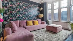 Ein Wohnzimmer mit einem rosa Ecksofa, einer runden Stehlampe und einem großen Fenster mit Blick auf Gebäude.