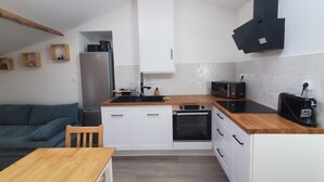 A compact kitchen with white cabinets, a wooden countertop, a built-in oven, a microwave, and a toaster. There is a wooden dining table and a green sofa in the adjacent living area.