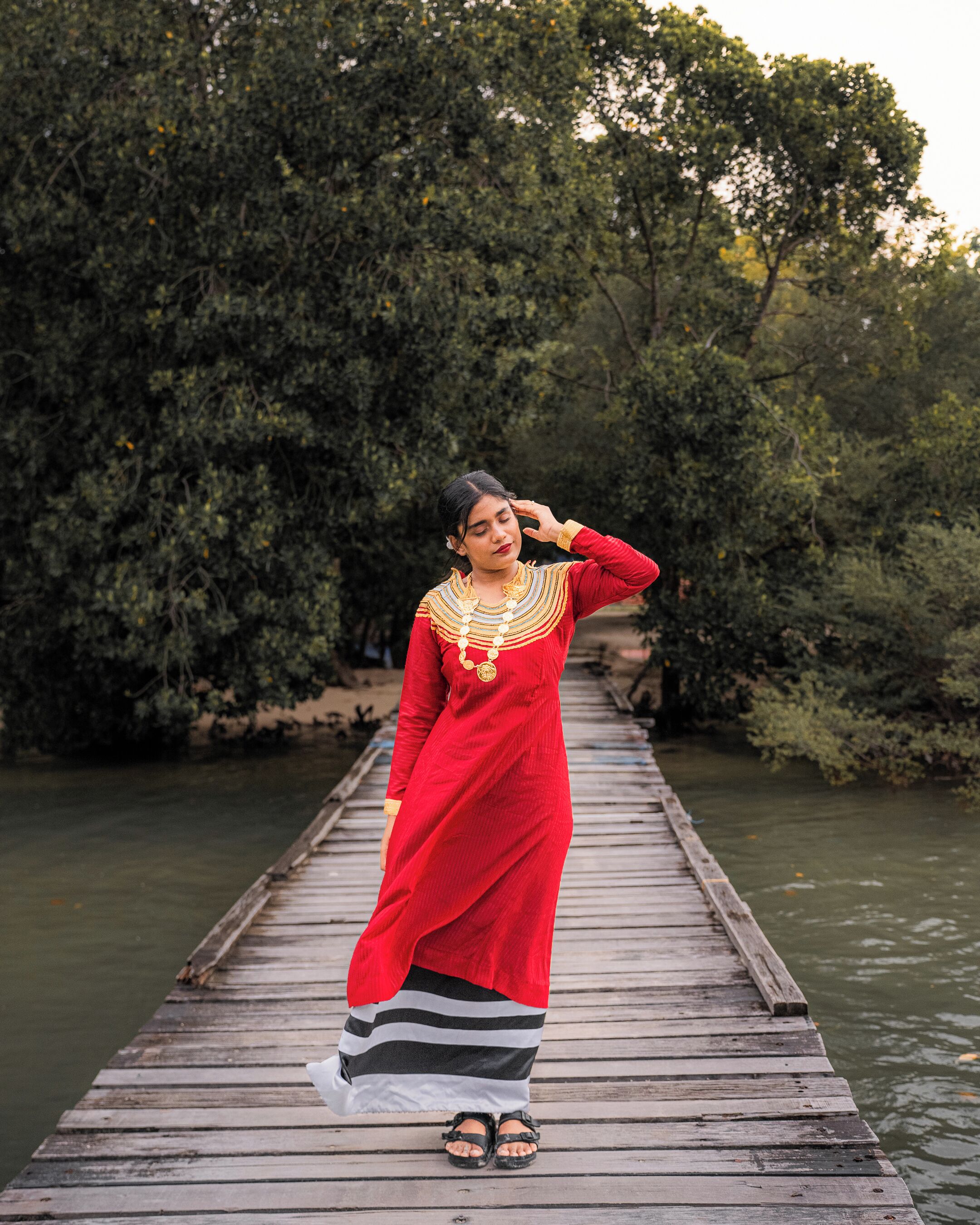 A person standing on a wooden bridge over water, wearing a red dress with gold jewelry.