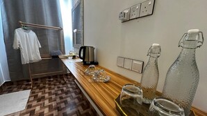 A hotel room with a wooden floor, a wooden counter with a kettle, glasses, and bottles, a hanging rack with a white shirt, and a window with curtains.