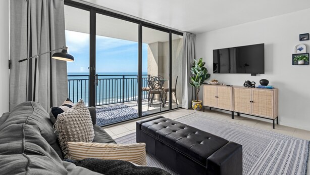 Ett modernt vardagsrum med en soffa, en svart pall, ett matbord med stolar, en tv som är monterad på väggen och utsikt över havet genom stora glasdörrar.