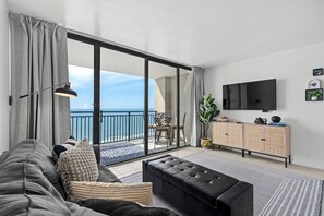 A modern living room with a sofa, a black ottoman, a dining table with chairs, a TV mounted on the wall, and a view of the sea through large glass doors. - Oceanfront Stay with Private Balcony (Myrtle Beach)