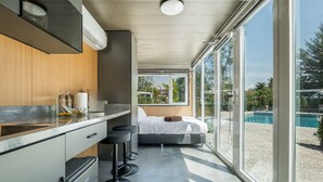 A modern kitchen with a built-in oven, a dining area with bar stools, a large window with a view of a pool, and a bedroom with a bed and a window. - Apple House (Choeng Thale)