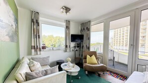 A hotel room with a sofa, armchair, and a bed. There is a small table with a vase and a book. A television is mounted on the wall. - Vacation apartment Watt`n Blick 220: Comfortable vacation right on the beach in Cuxhaven (Cuxhaven)