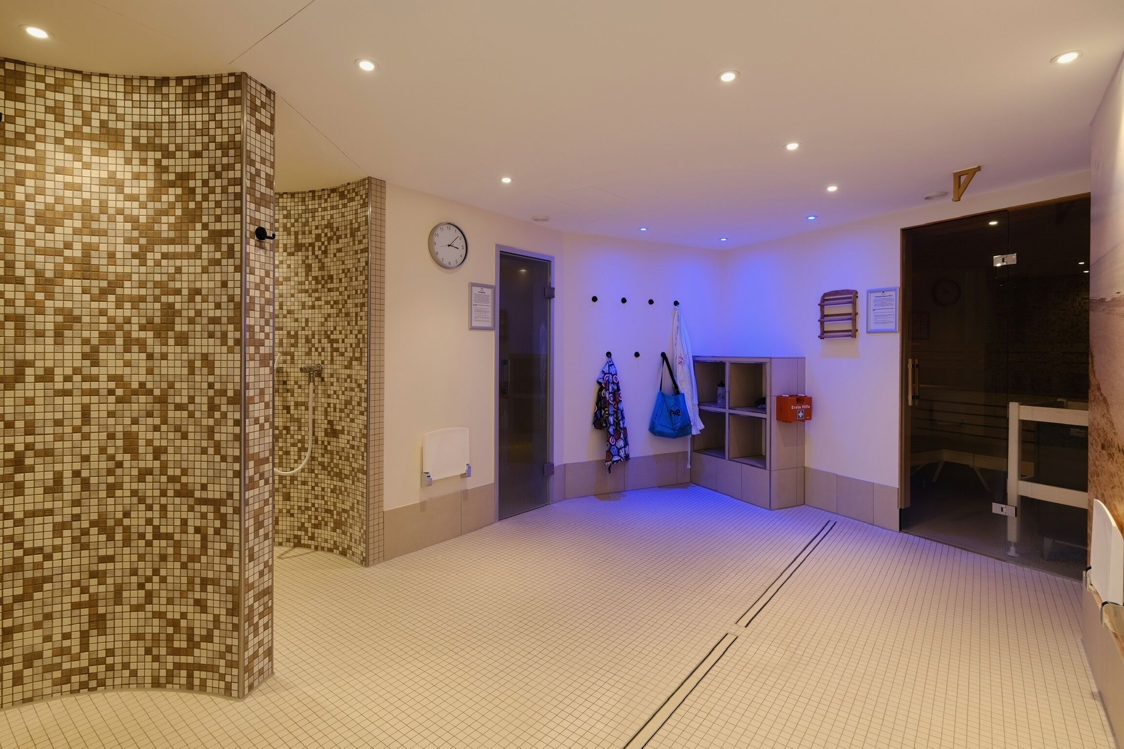 Eine Hotelhalle mit Fliesenboden, einer Uhr an der Wand und einem Kleiderständer mit blauer Tasche.