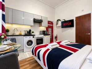 A modern hotel room with a kitchen, a bed with a red, white, and blue striped bedspread, and a television displaying 'NETFLIX'.