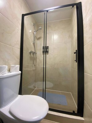 A shower stall with a glass door, a toilet with a paper towel holder, and a blue mat on the floor.