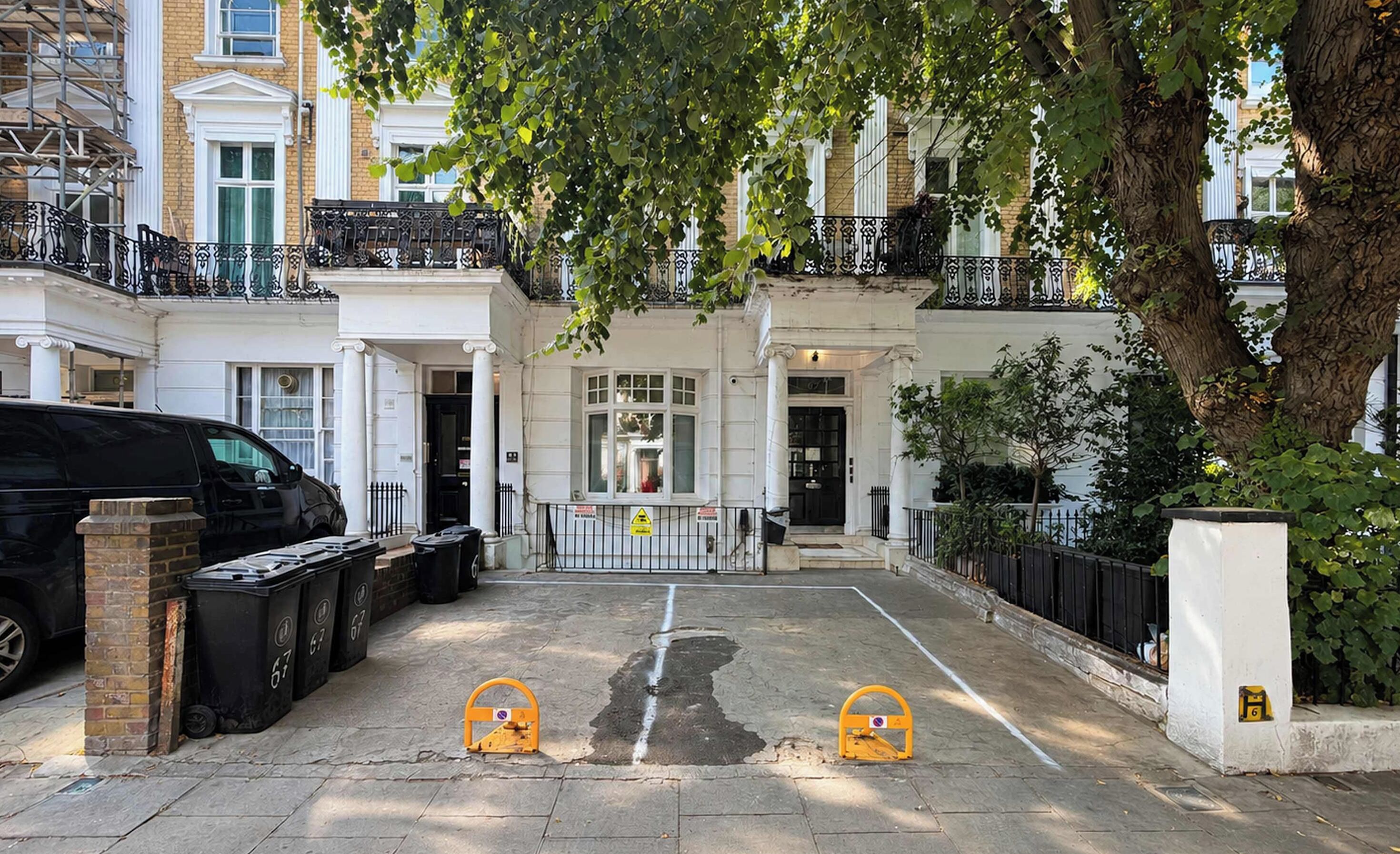 Une rue résidentielle avec une voiture noire garée sur le côté gauche, un arbre au premier plan et un bâtiment blanc avec un portail noir à l’arrière-plan.
