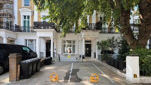 A residential street with a black car parked on the left, a tree in the foreground, and a white building with a black gate in the background. - London Style (5) Apartment (Greater London)