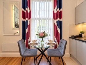 A dining area with a glass table, two grey chairs, a vase with flowers, and a window with blinds. - London Style (5) Apartment (Greater London)