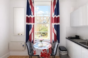Eine Küche mit Fenster, einem Tisch mit Blumenmuster-Tischdecke und Blick auf einen Garten.