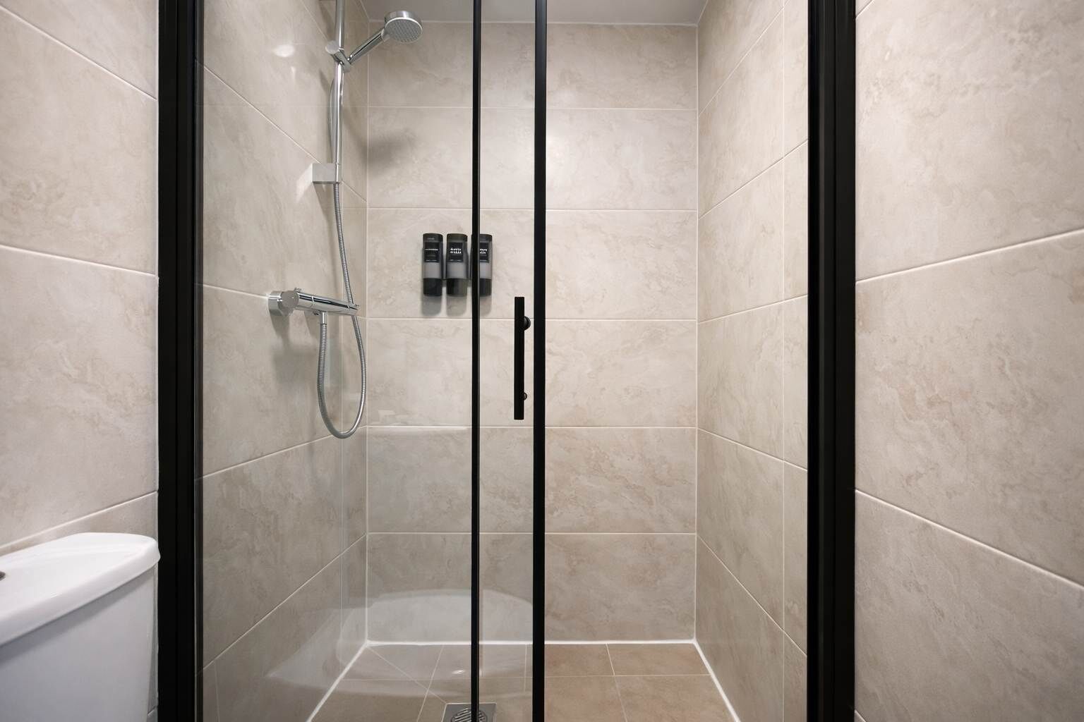 Une douche en verre avec un pommeau de douche à main et un robinet mural, des toilettes et un ensemble de produits de toilette fixés au mur.