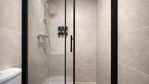 A glass-enclosed shower with a handheld showerhead and a wall-mounted faucet, a toilet, and a set of toiletries on the wall.