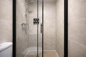 Ducha envuelta en cristal con cabezal de ducha manual y llave empotrada en la pared, inodoro y un juego de artículos de aseo colgados en la pared.
