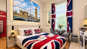 A hotel room with a large bed, a painting of London, a red telephone booth, and a view of greenery outside. - London Style (5) Apartment (Greater London)