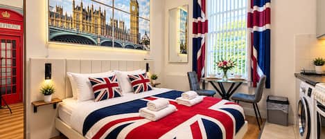 A hotel room with a large bed, a painting of London, a red telephone booth, and a view of greenery outside.