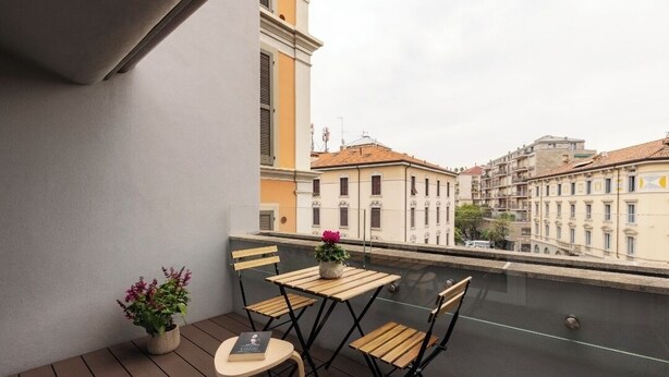 A balcony with a wooden table and chairs, a potted plant, and a view of buildings across a water body. - DONIZETTI STUDIOS (Bergamo)