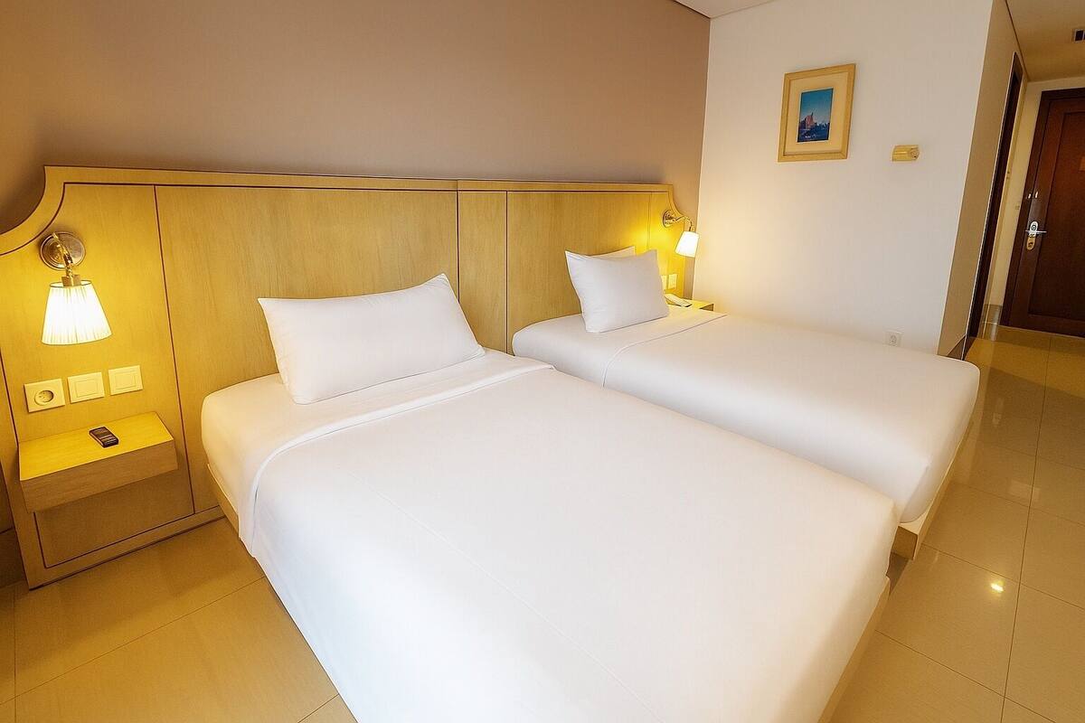 A hotel room with two beds, a wooden headboard, bedside lamps, a nightstand, and a framed picture on the wall.