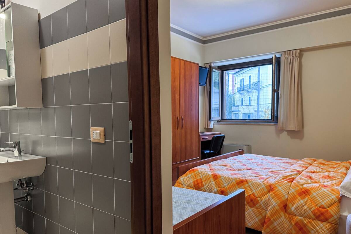 Ein Zimmer mit Bett, Schreibtisch, einem Fenster mit Vorhängen sowie einem Badezimmer mit Waschbecken und Spiegel.