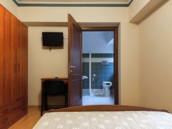 A bedroom with a bed, a wooden wardrobe, a desk with a chair, a television on the wall, and a bathroom visible through an open door. - Hotel Fanesia Coliving (Fano Adriano)