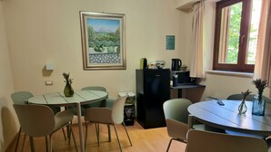 A dining area with a round table, chairs, a refrigerator, a coffee machine, and a window with curtains. - Hotel Fanesia Coliving (Fano Adriano)